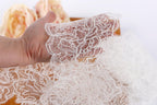 A hand is holding a piece of white lace with a floral pattern, against a blurred background of more lace pieces.