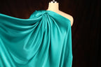 A mannequin wearing a flowing, turquoise satin garment with a draped, one-shoulder design.