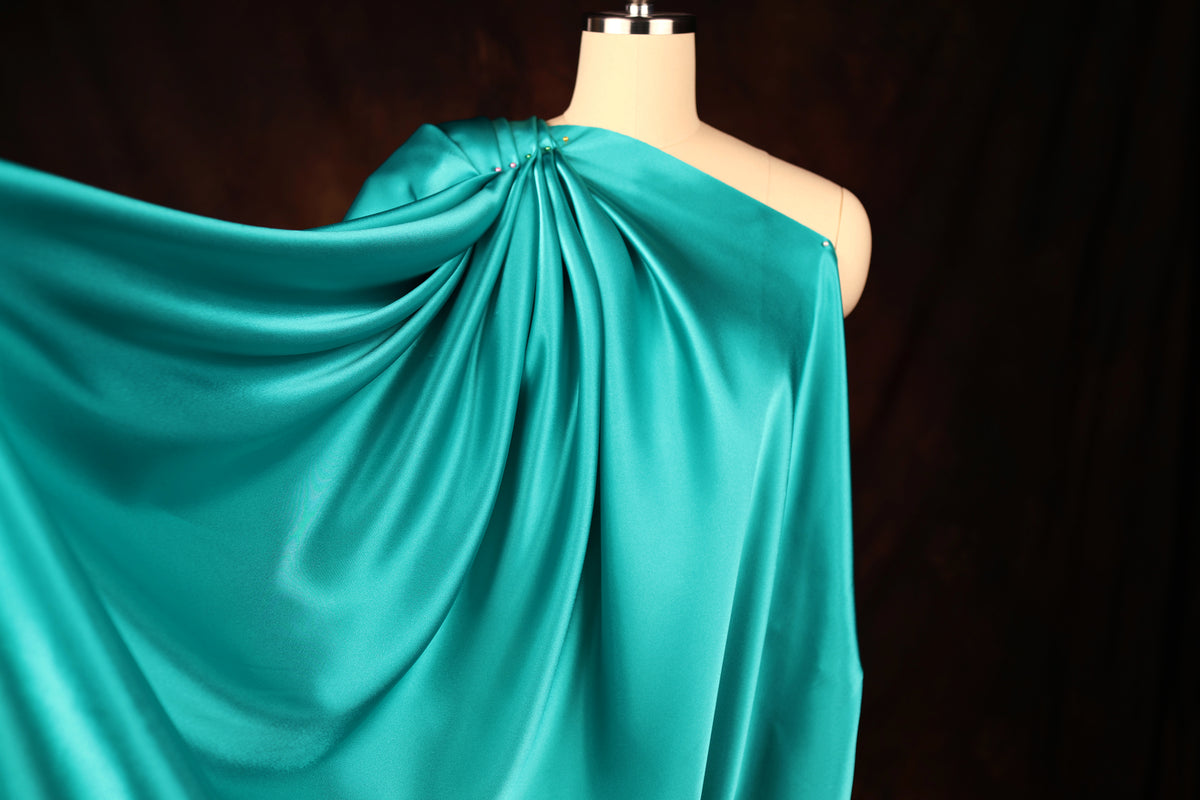 A mannequin wearing a flowing, turquoise satin garment with a draped, one-shoulder design.
