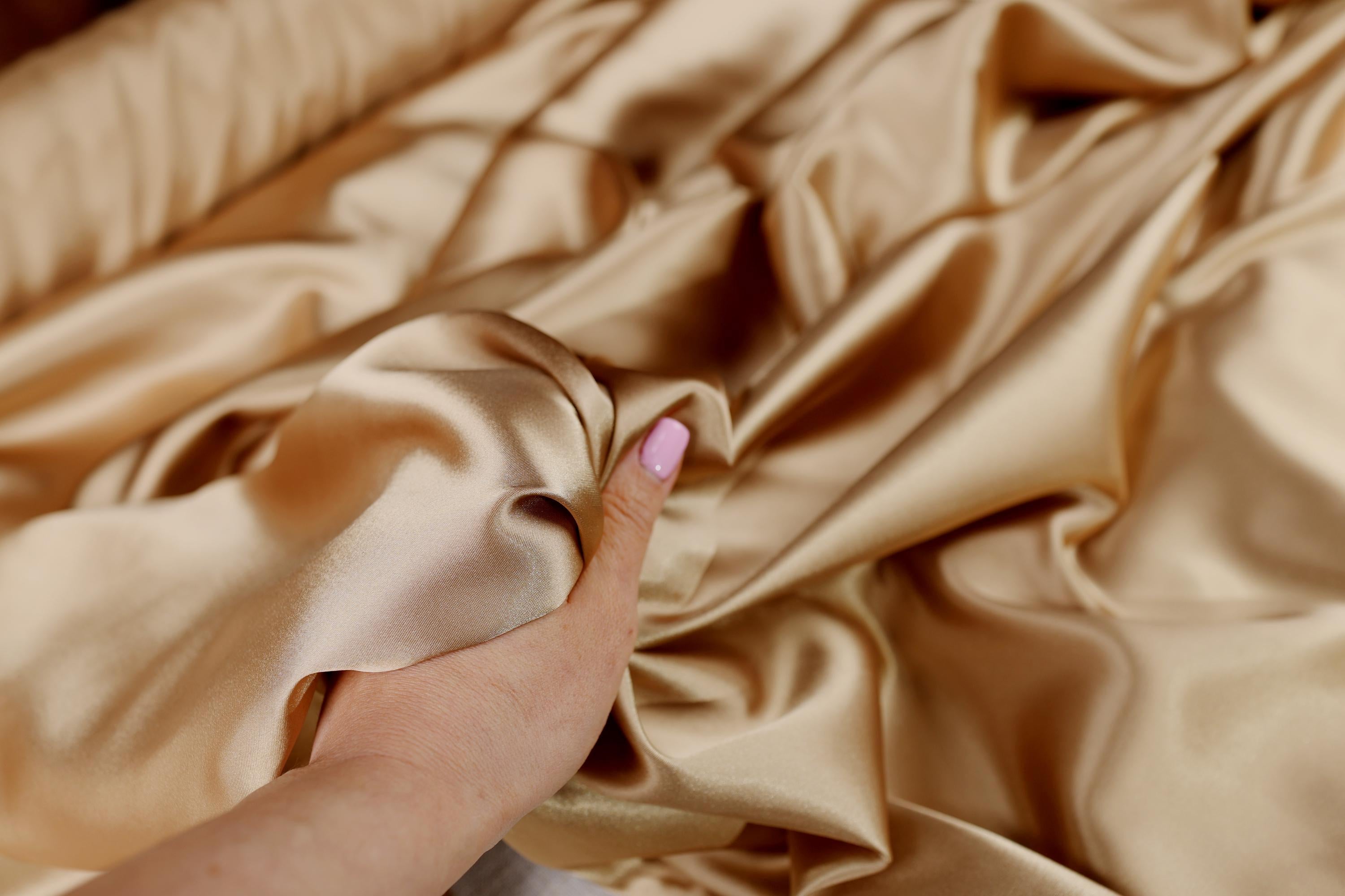 A hand is holding a piece of gold satin fabric, which appears to be soft and smooth.