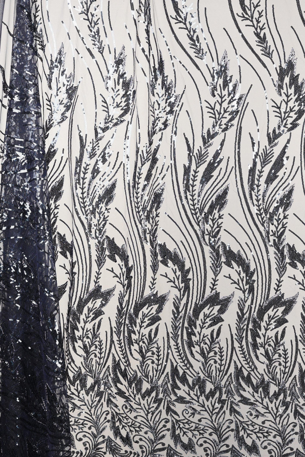 a close-up view of a fabric with a repeating pattern of black and white floral designs. The pattern consists of various leaf-like shapes and lines, creating a visually striking and intricate design. The fabric appears to be made of a material with a shimmering or sequined effect, adding a touch of elegance and sophistication to the overall appearance.