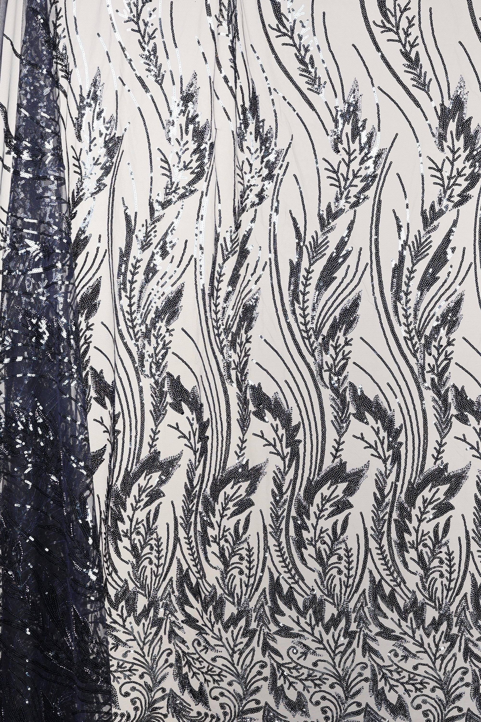 a close-up view of a fabric with a repeating pattern of black and white floral designs. The pattern consists of various leaf-like shapes and lines, creating a visually striking and intricate design. The fabric appears to be made of a material with a shimmering or sequined effect, adding a touch of elegance and sophistication to the overall appearance.