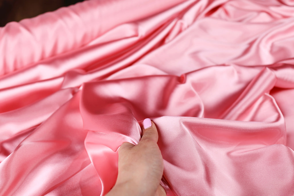 A hand is holding a piece of pink satin fabric, which appears to be soft and smooth.