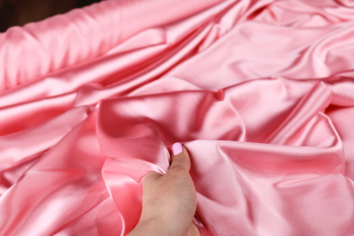 A hand is holding a piece of pink satin fabric, which appears to be soft and smooth.