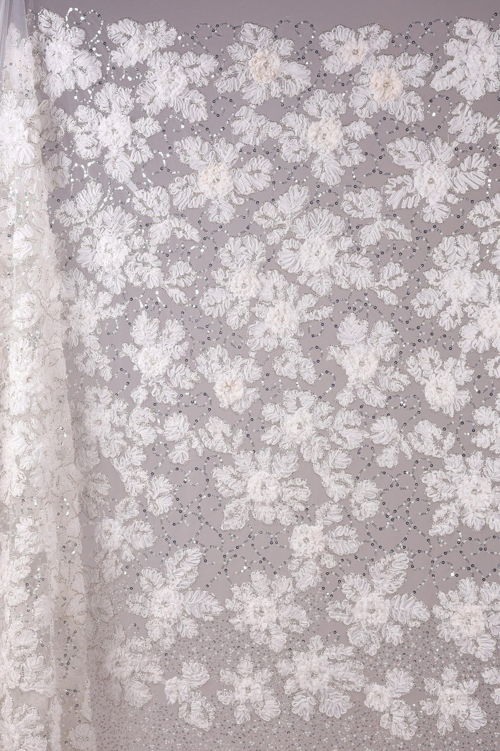 a close-up view of a delicate, white lace fabric adorned with a floral pattern. The fabric is intricately designed with numerous small white flowers and leaves, creating a visually appealing and delicate appearance. The close-up perspective allows for a detailed examination of the fabric's texture and design.
