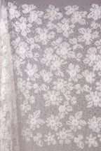 a close-up view of a delicate, white lace fabric adorned with a floral pattern. The fabric is intricately designed with numerous small white flowers and leaves, creating a visually appealing and delicate appearance. The close-up perspective allows for a detailed examination of the fabric's texture and design.