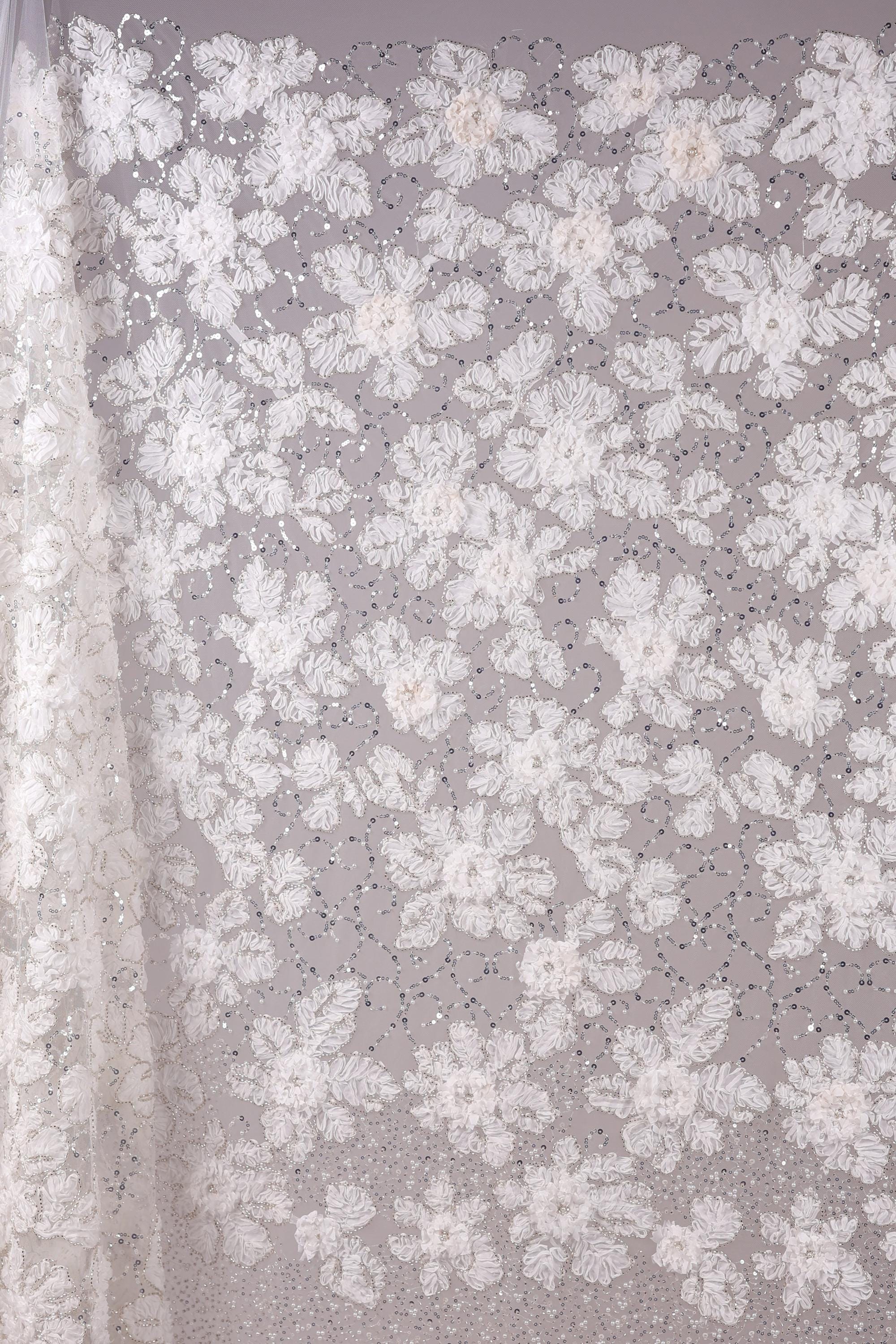 a close-up view of a delicate, white lace fabric adorned with a floral pattern. The fabric is intricately designed with numerous small white flowers and leaves, creating a visually appealing and delicate appearance. The close-up perspective allows for a detailed examination of the fabric's texture and design.