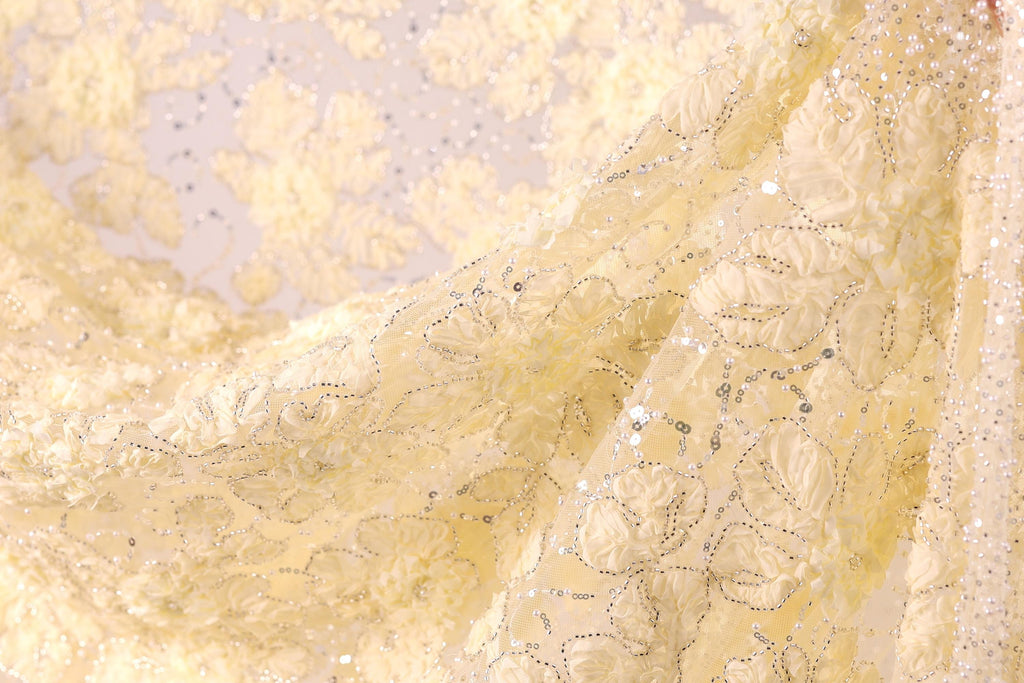 a close-up view of a delicate, yellow fabric with a floral pattern. The fabric appears to be made of a soft, shiny material and has a subtle, sparkling texture. The background is blurred, drawing focus to the fabric and its intricate design.