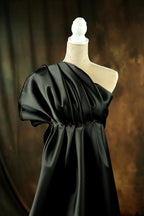 A black one-shoulder dress displayed on a mannequin, with a dark background.