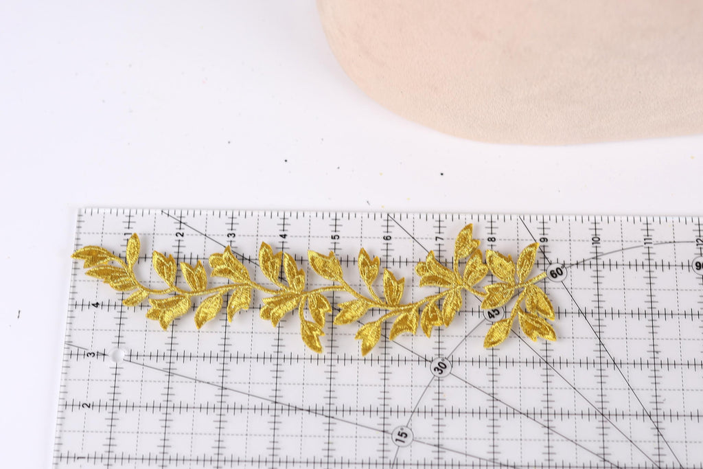 a close-up view of a gold embroidered leaf design placed on a ruler, indicating that it is being measured or sized for a sewing or crafting project.