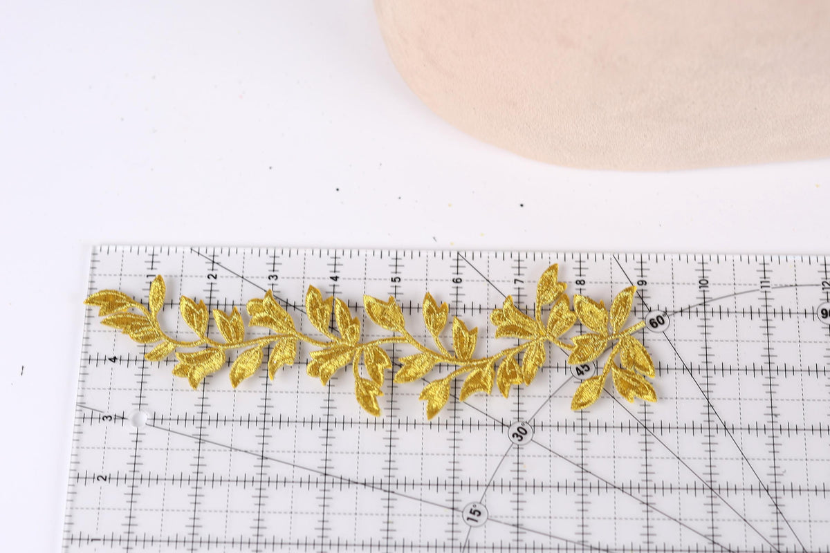 a close-up view of a gold embroidered leaf design placed on a ruler, indicating that it is being measured or sized for a sewing or crafting project.