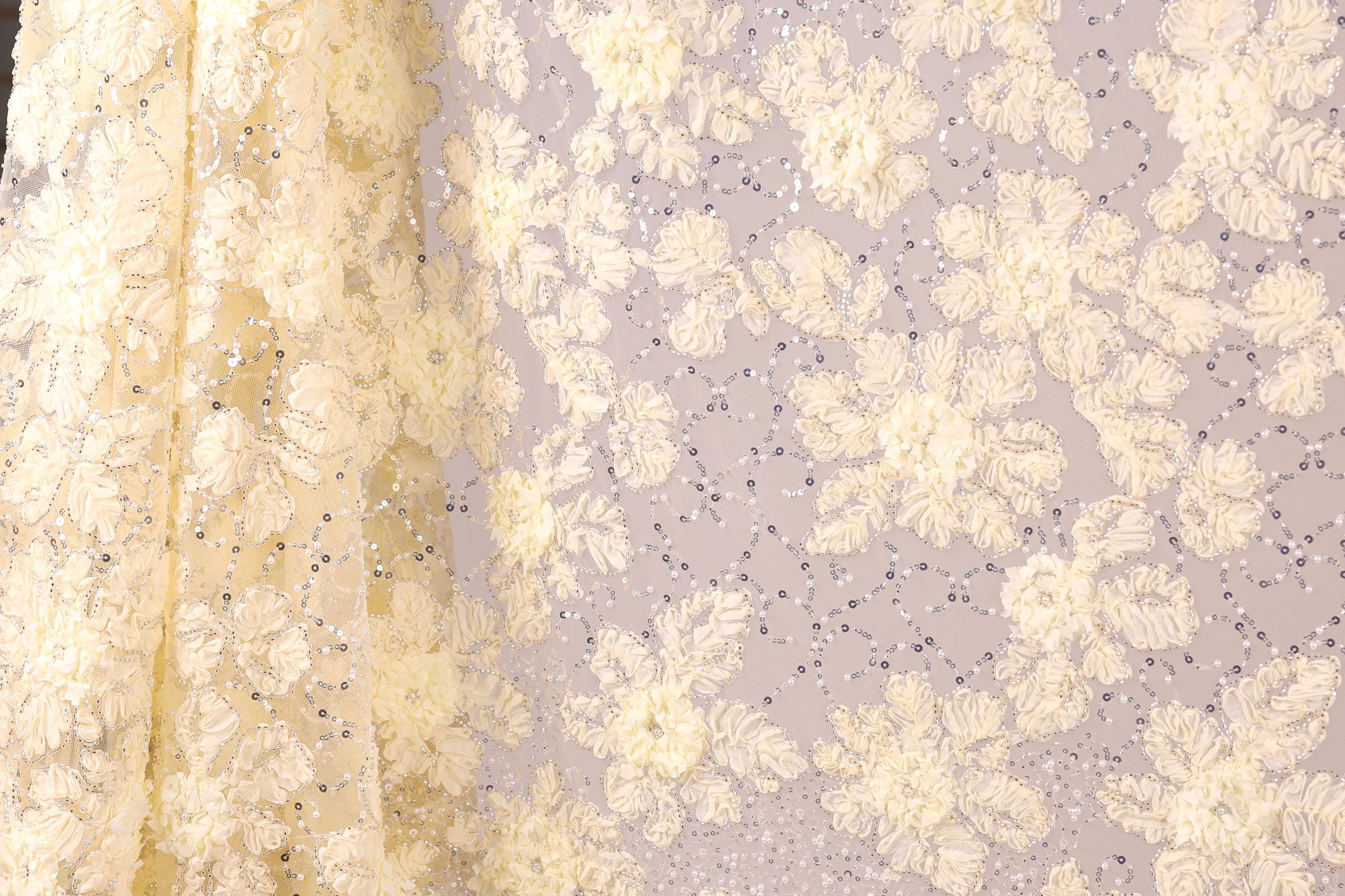 a close-up view of a fabric with a floral pattern, including white and yellow flowers and sequins. The fabric appears to be made of a sheer material, giving it a delicate and elegant appearance.