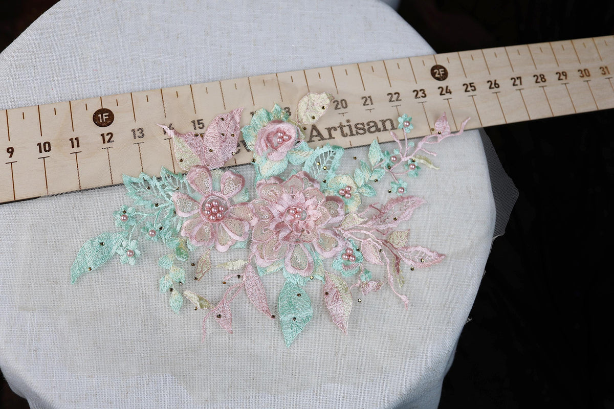 a close-up view of a delicate floral embroidery piece with pink and green flowers, placed on a white surface. A ruler is visible in the background, likely used for measuring the size of the embroidery.