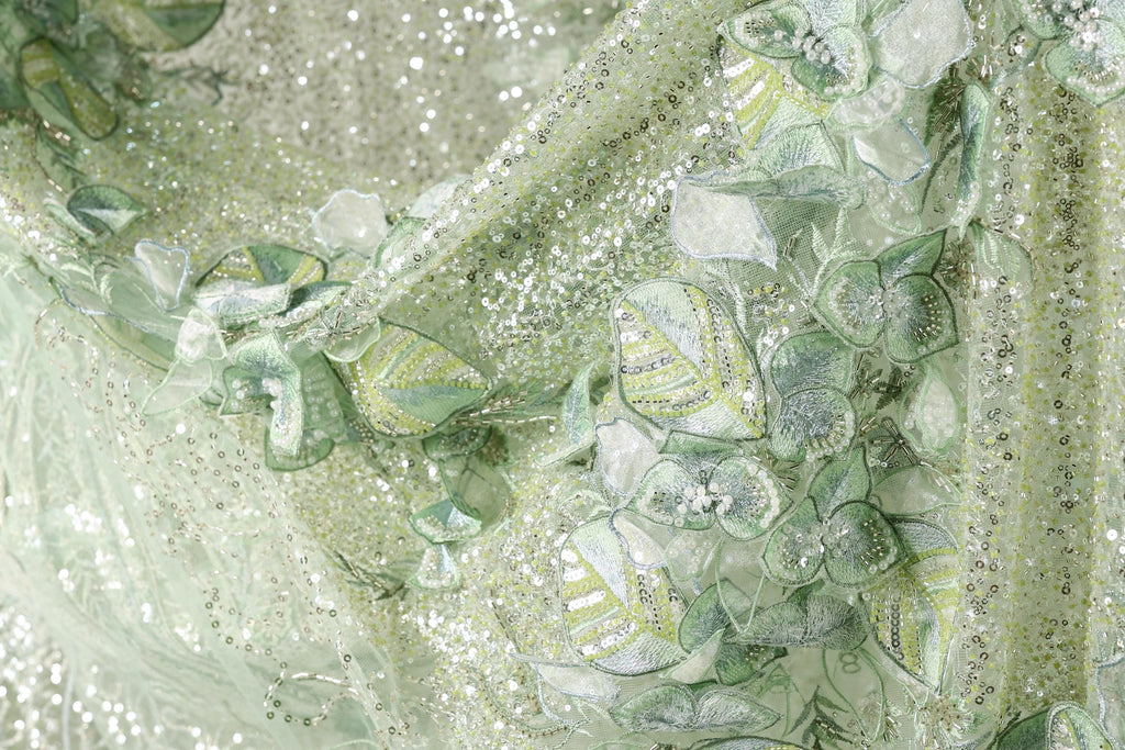 The image is a close-up of a fabric with a floral pattern, featuring green and white colors and small sequins or beads.