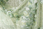 The image is a close-up of a fabric with a floral pattern, featuring green and white colors and small sequins or beads.