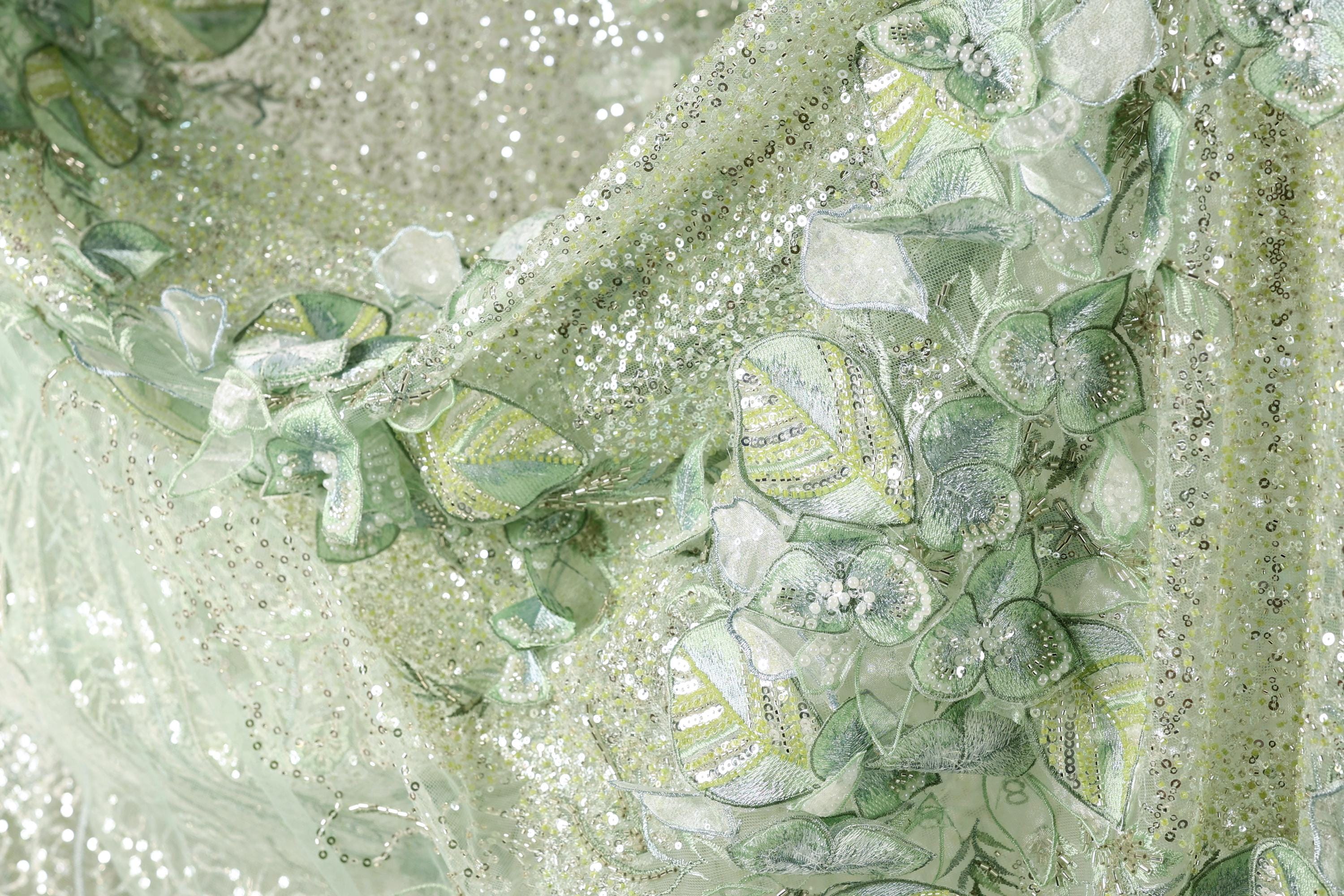 The image is a close-up of a fabric with a floral pattern, featuring green and white colors and small sequins or beads.