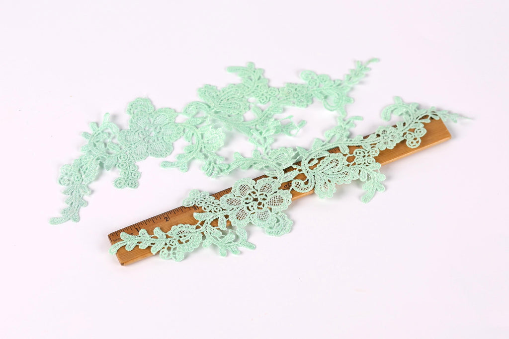 a collection of small, delicate green lace pieces arranged on a wooden ruler, which is placed on a white background.