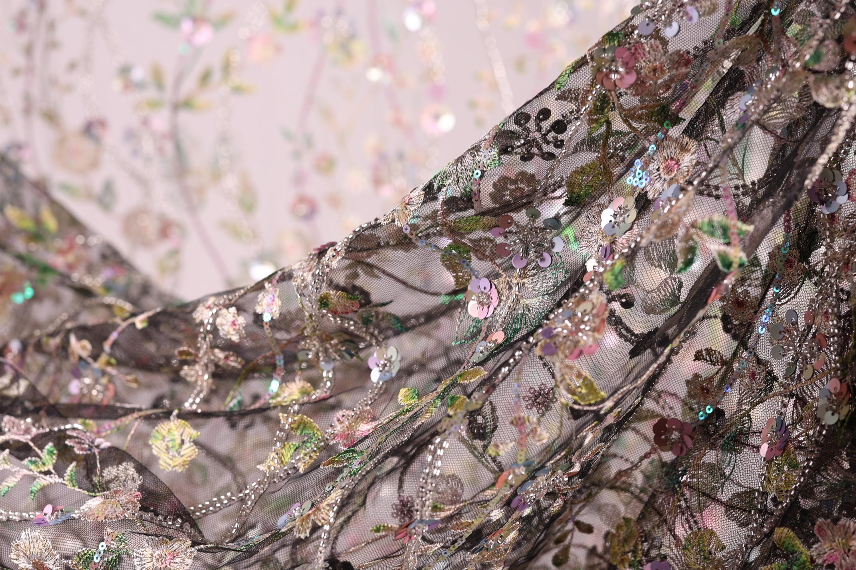 a close-up view of a delicate, intricately embroidered fabric with a floral pattern. The fabric appears to be made of a sheer material, with a variety of colors and designs, including pink, green, and black elements. The image is a close-up of the fabric, focusing on the details and textures of the embroidery.