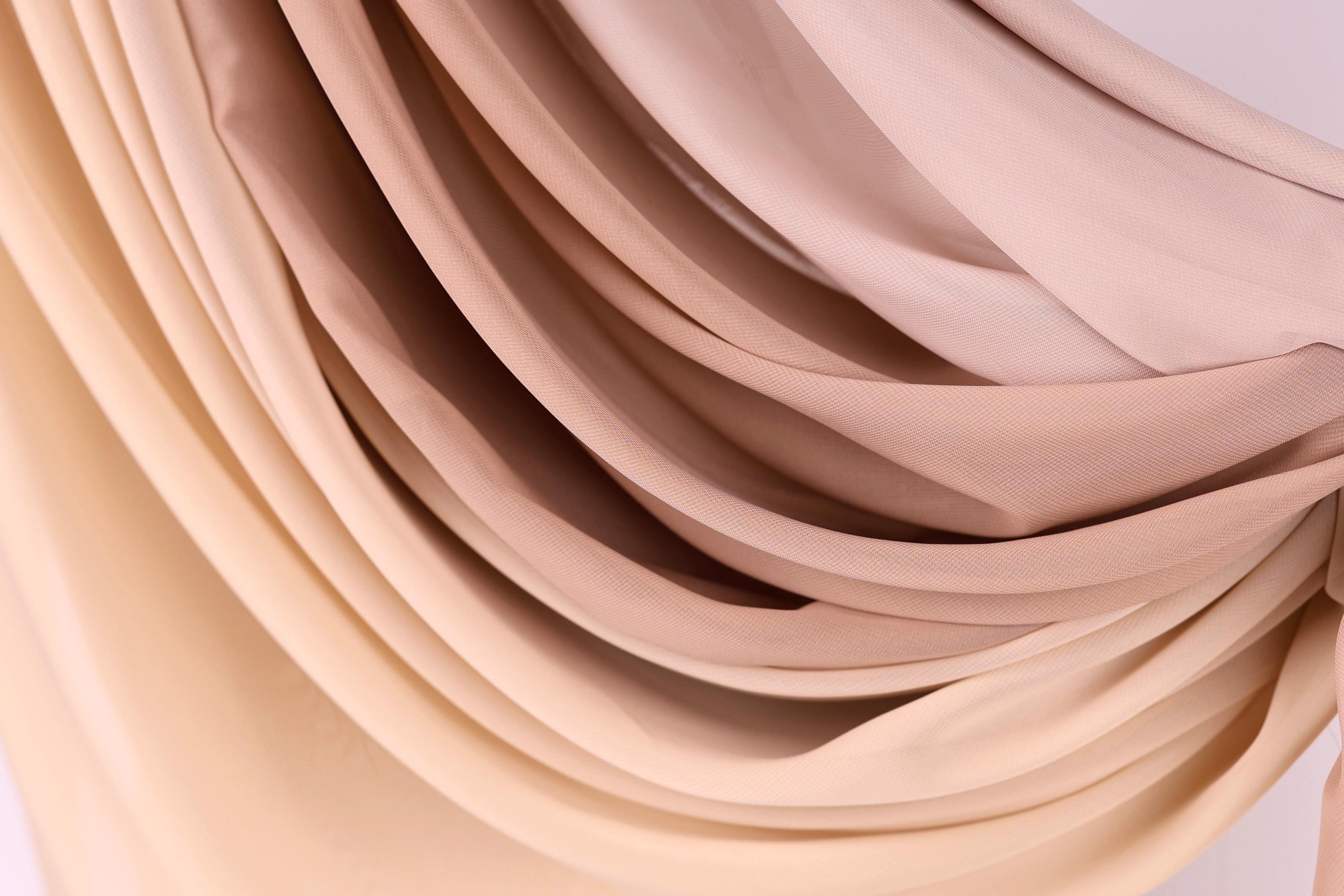 a close-up view of a fabric with a gradient of colors, transitioning from a light beige to a darker beige. The fabric appears to be made of a soft, flowing material, possibly silk or chiffon. The colors and texture of the fabric create a visually appealing and textured appearance.