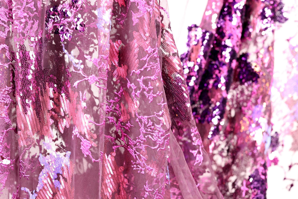 a close-up view of a fabric with a vibrant, multicolored pattern of pink and purple sequins. The fabric appears to be a sheer material, with a delicate and intricate design. The background is blurred, drawing focus to the fabric and its details.