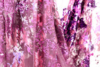 a close-up view of a fabric with a vibrant, multicolored pattern of pink and purple sequins. The fabric appears to be a sheer material, with a delicate and intricate design. The background is blurred, drawing focus to the fabric and its details.