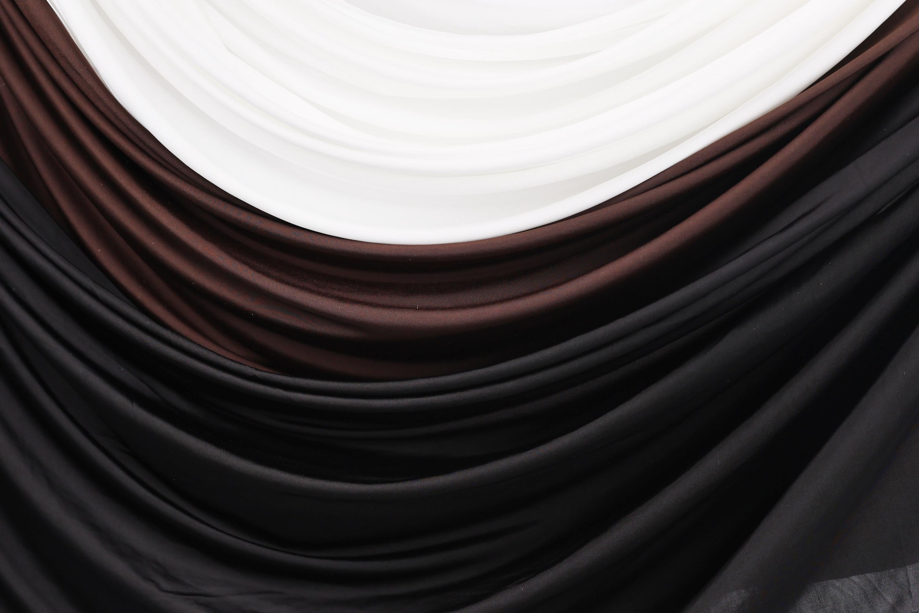 a close-up view of a fabric with a gradient of colors, including black, brown, and white. The fabric appears to be made of a soft, flowing material, possibly silk or chiffon. The colors are layered and interwoven, creating a visually appealing and textured appearance.