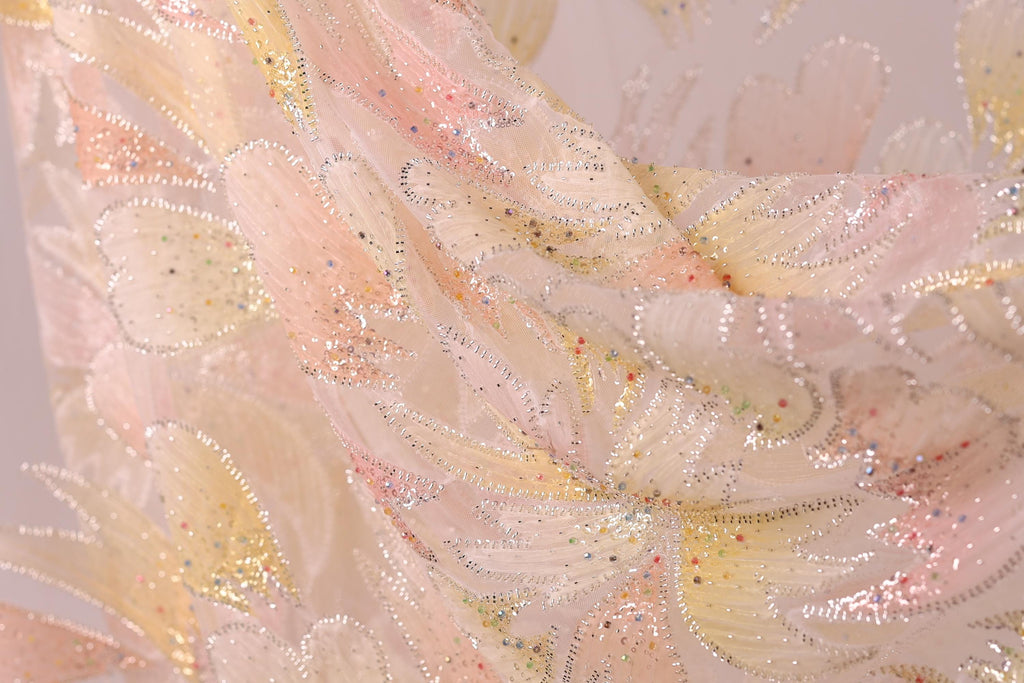 The image is a close-up of a delicate, intricately designed fabric with a floral pattern. The fabric appears to be made of a soft, light-colored material, possibly silk or chiffon, with a shimmering, sparkly texture. The pattern consists of numerous small, overlapping flowers or leaves, creating a visually striking and detailed design.