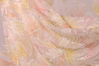 The image is a close-up of a delicate, intricately designed fabric with a floral pattern. The fabric appears to be made of a soft, light-colored material, possibly silk or chiffon, with a shimmering, sparkly texture. The pattern consists of numerous small, overlapping flowers or leaves, creating a visually striking and detailed design.