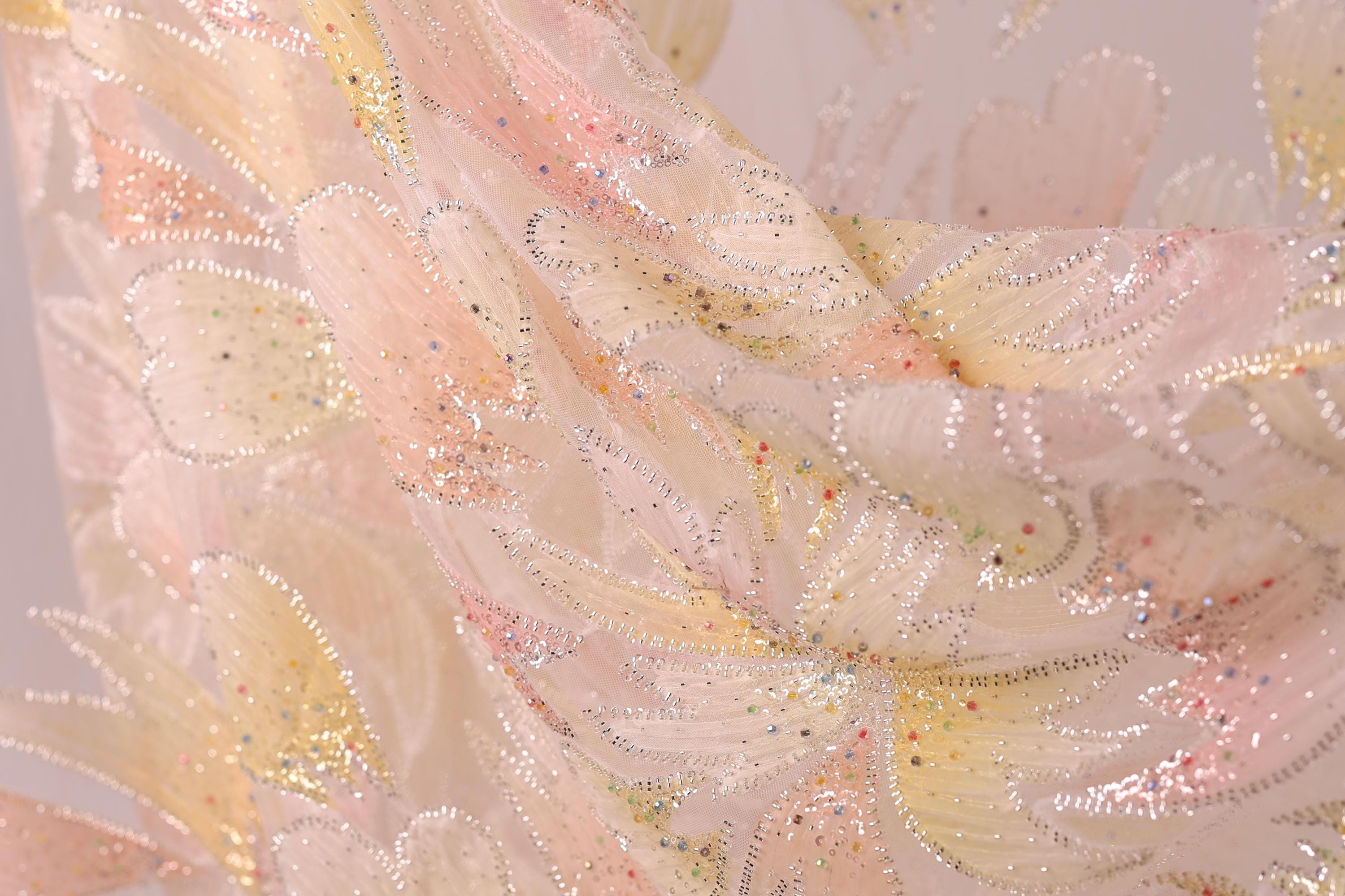 The image is a close-up of a delicate, intricately designed fabric with a floral pattern. The fabric appears to be made of a soft, light-colored material, possibly silk or chiffon, with a shimmering, sparkly texture. The pattern consists of numerous small, overlapping flowers or leaves, creating a visually striking and detailed design.