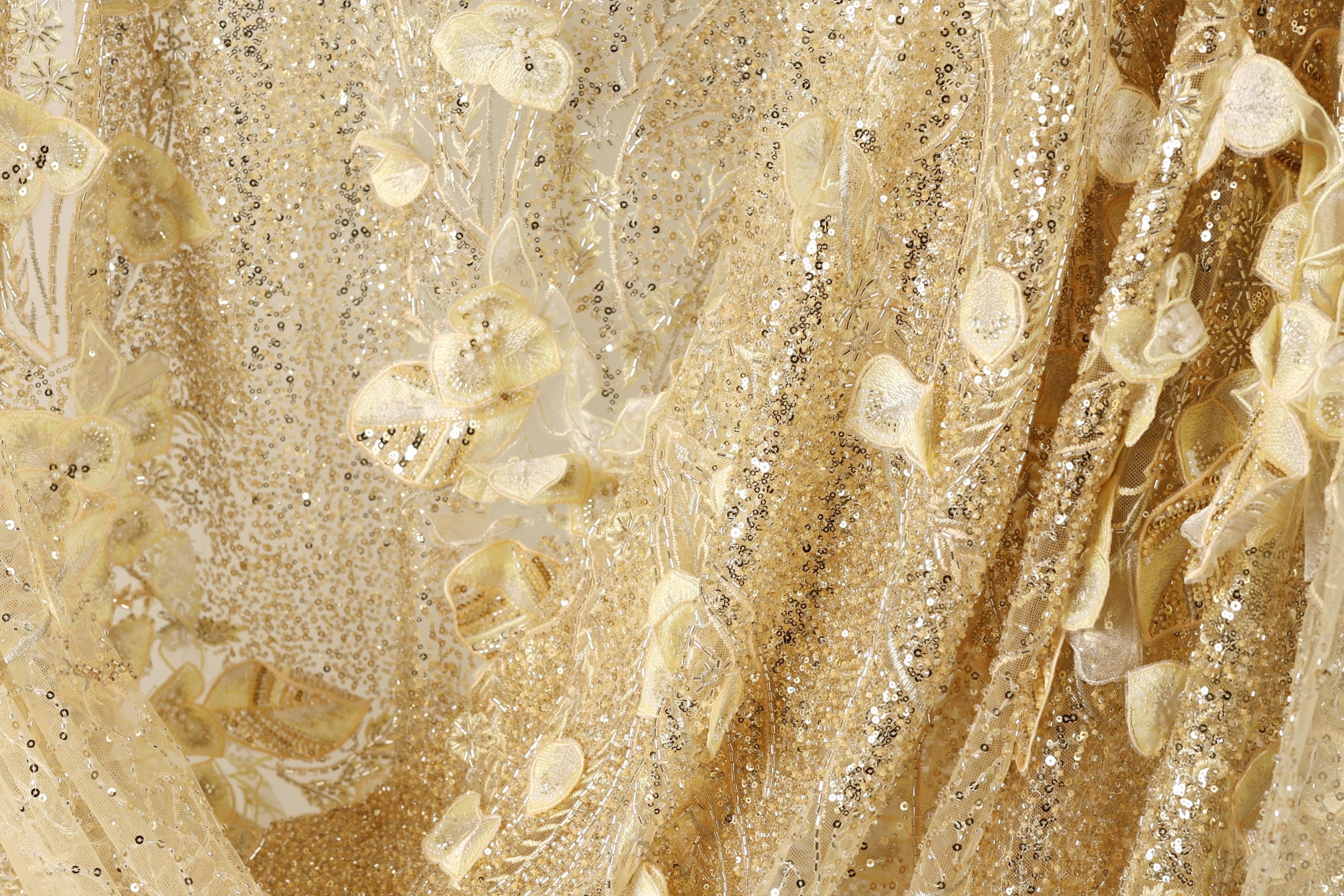The image is a close-up of a fabric with a gold and white color scheme, featuring a floral pattern and sequin accents.