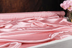 a close-up view of a soft, pink satin fabric draped over a surface, with a vase of pink flowers visible in the background.