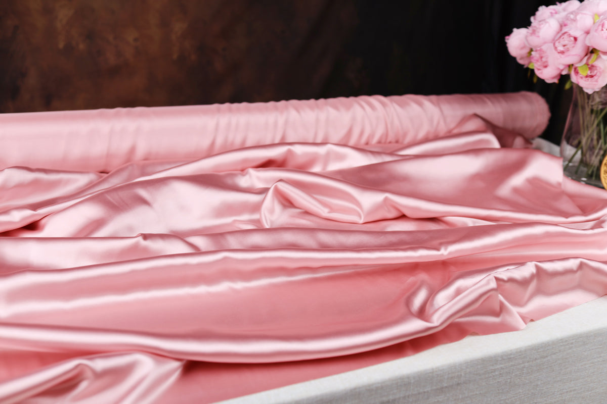 a close-up view of a soft, pink satin fabric draped over a surface, with a vase of pink flowers visible in the background.