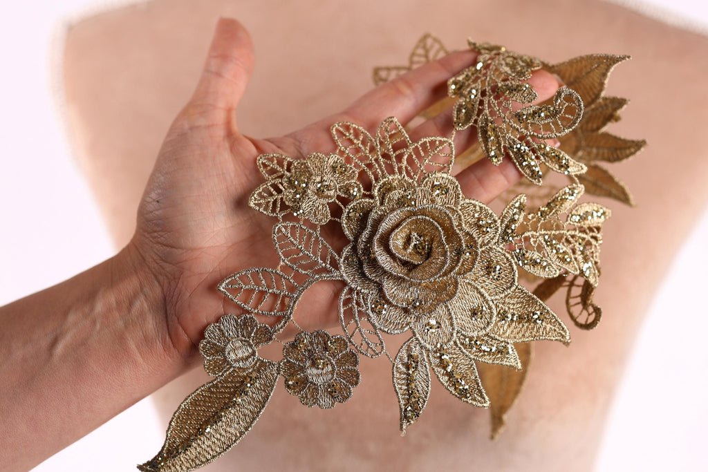 A hand holding a piece of gold embroidered jewelry, featuring a large rose and various leaf-like elements.