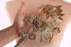 A hand holding a piece of gold embroidered jewelry, featuring a large rose and various leaf-like elements.