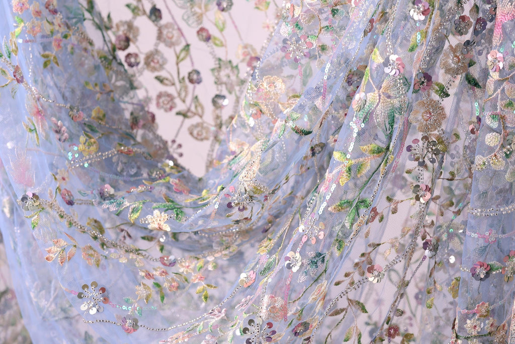 The image is a close-up view of a delicate, intricately embroidered fabric with a floral pattern. The fabric appears to be made of sheer material, giving it a soft and delicate appearance. The embroidery features a variety of flowers and leaves in different colors, creating a visually appealing and intricate design.