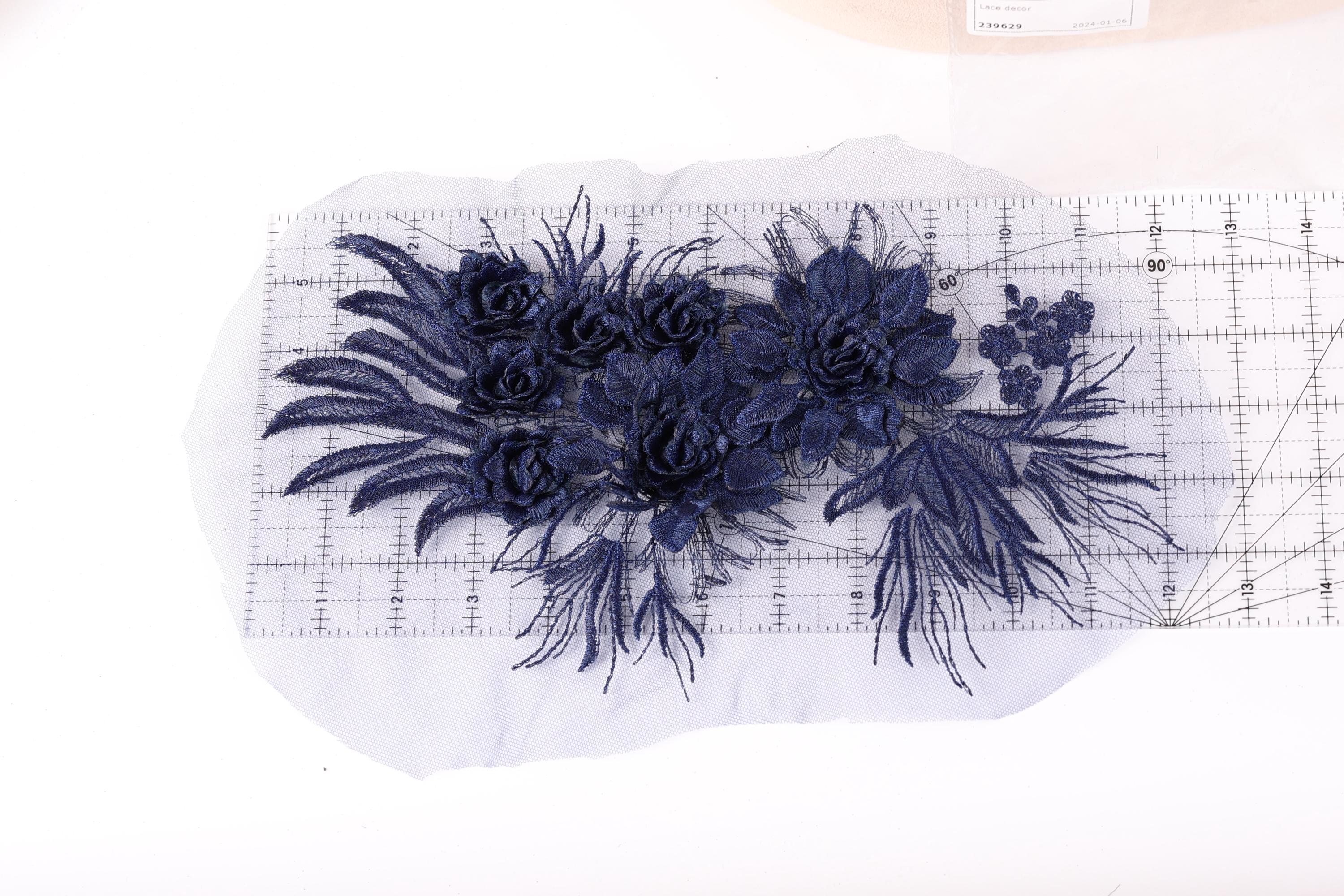 a close-up view of a piece of fabric with a floral design, including blue flowers and leaves. The fabric appears to be a lace or a similar type of material, and it is placed on a white surface. There is a ruler visible in the image, which is likely used for measuring or cutting the fabric.