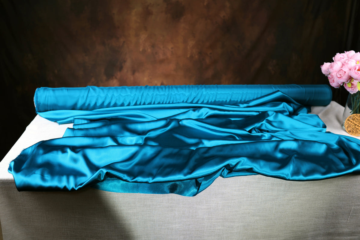 a roll of blue satin fabric on a table, with a vase of pink flowers placed next to it.
