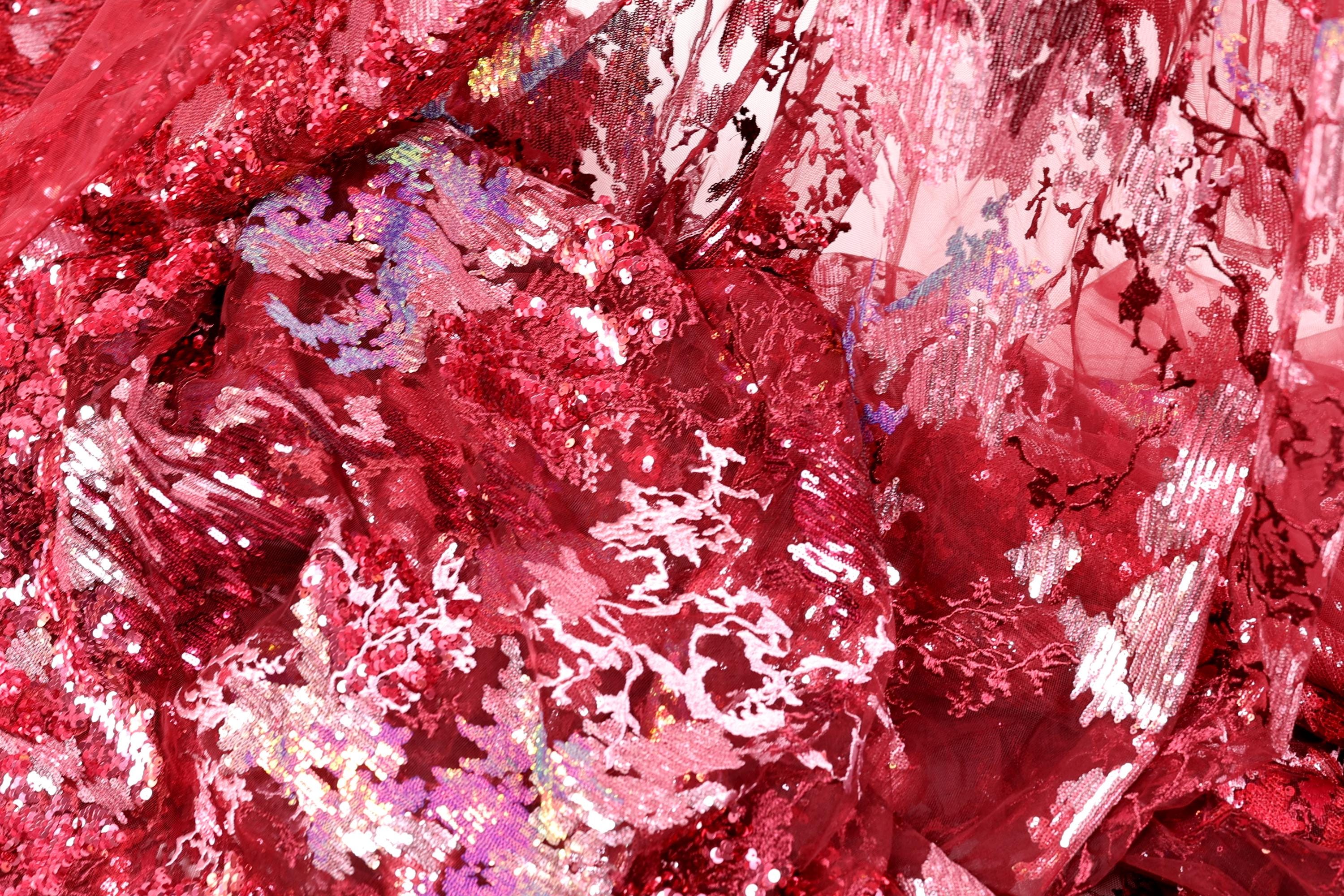 The image is a close-up of a fabric with a red and pink color scheme, featuring a variety of textures and patterns, including sequins and lace-like elements.