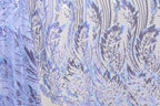a close-up view of a fabric with a repeating pattern of purple and white sequin designs. The pattern appears to be a floral or leaf-like design, with the sequins adding a touch of sparkle and texture to the fabric. The fabric is predominantly white, with the purple and white sequins creating a visually striking contrast. The image does not contain any text or other discernible objects, and the focus is solely on the intricate and detailed pattern of the fabric.