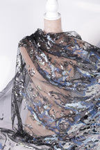 a close-up view of a piece of sheer, black, and blue fabric with a floral pattern. The fabric appears to be made of sequin material and is being held up by a white pole.