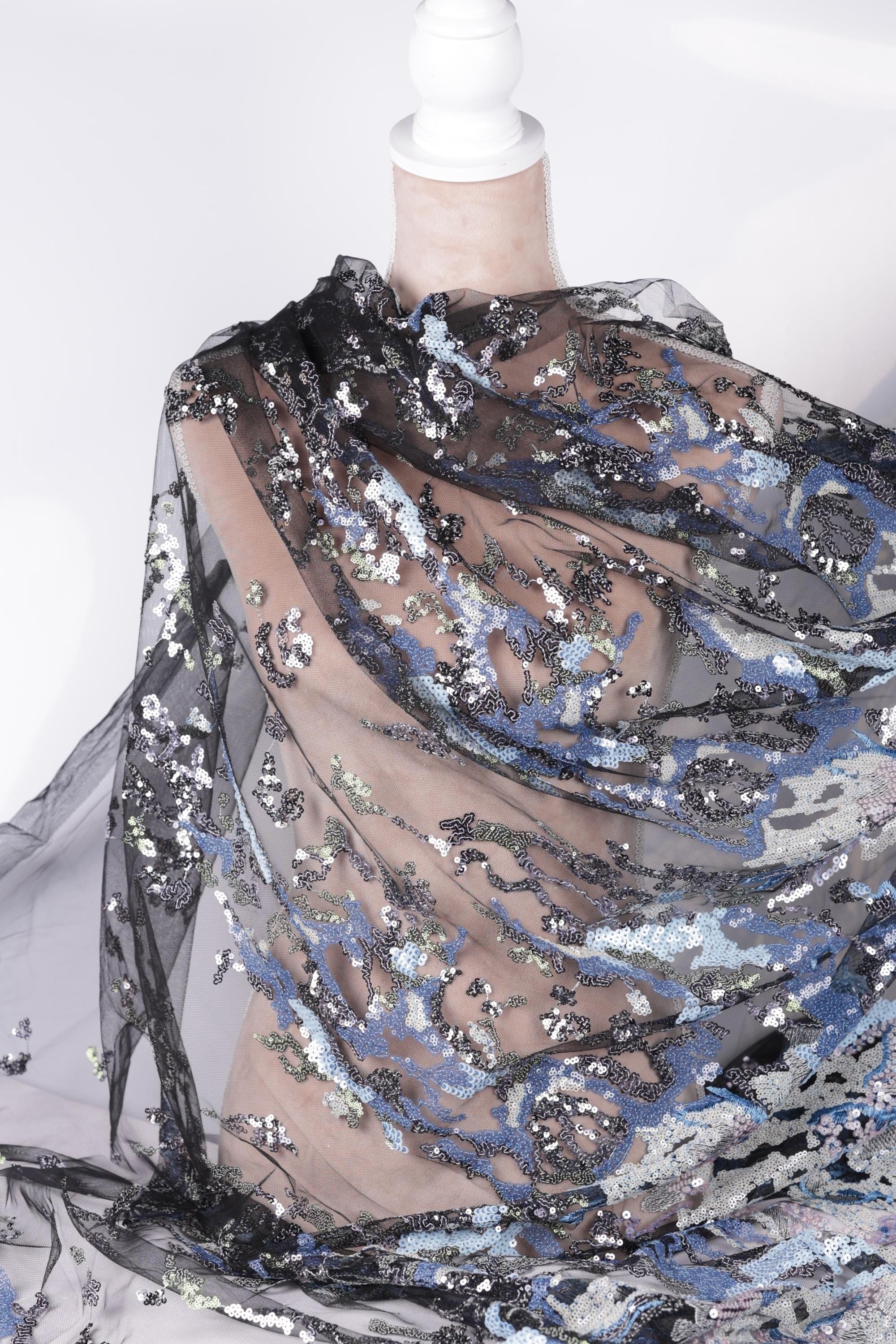 a close-up view of a piece of sheer, black, and blue fabric with a floral pattern. The fabric appears to be made of sequin material and is being held up by a white pole.