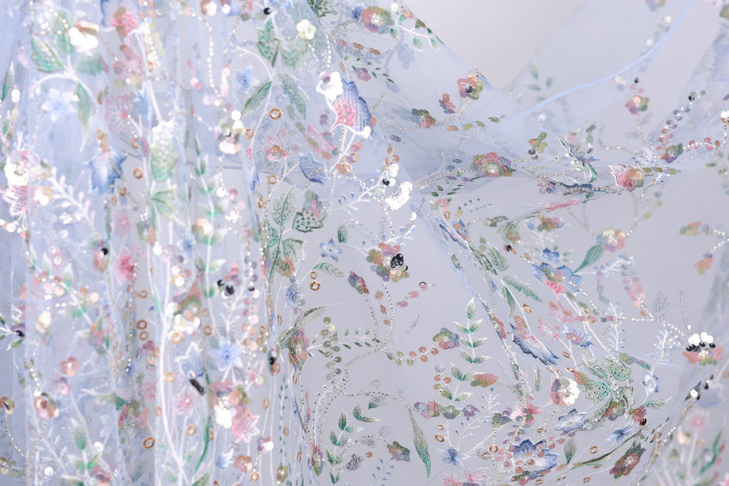 The image is a close-up view of a delicate, intricately embroidered fabric with a floral pattern. The fabric appears to be made of sheer material, allowing the colorful flowers and leaves to be clearly visible. The image does not contain any text or other discernible objects, and the focus is solely on the detailed embroidered design.