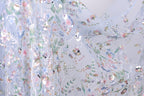 The image is a close-up view of a delicate, intricately embroidered fabric with a floral pattern. The fabric appears to be made of sheer material, allowing the colorful flowers and leaves to be clearly visible. The image does not contain any text or other discernible objects, and the focus is solely on the detailed embroidered design.