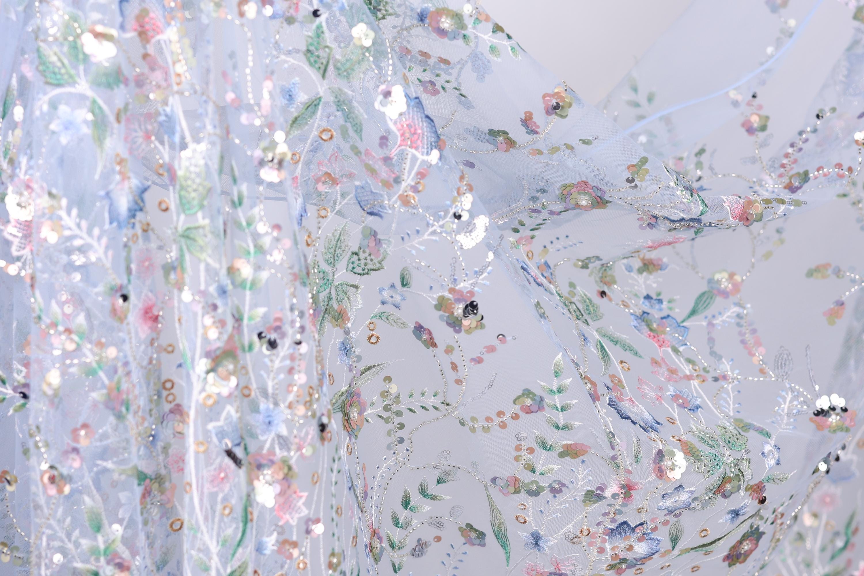 The image is a close-up view of a delicate, intricately embroidered fabric with a floral pattern. The fabric appears to be made of sheer material, allowing the colorful flowers and leaves to be clearly visible. The image does not contain any text or other discernible objects, and the focus is solely on the detailed embroidered design.