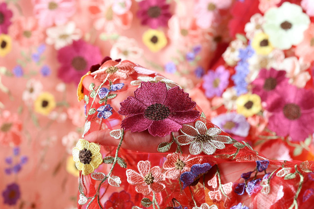 a close-up view of a fabric with a vibrant floral pattern, including red, pink, yellow, and purple flowers. The fabric appears to be made of a sheer material, with the flowers intricately embroidered or sewn onto it. The background is blurred, drawing focus to the colorful floral design in the foreground.