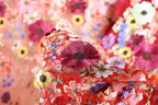 a close-up view of a fabric with a vibrant floral pattern, including red, pink, yellow, and purple flowers. The fabric appears to be made of a sheer material, with the flowers intricately embroidered or sewn onto it. The background is blurred, drawing focus to the colorful floral design in the foreground.