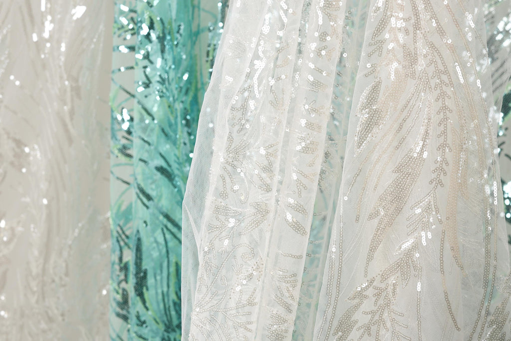 a close-up view of a white fabric with a floral pattern, as well as a fabric with a blue and green floral pattern. The fabrics appear to be made of sequins or other embellishments, adding a touch of sparkle and color to the scene.