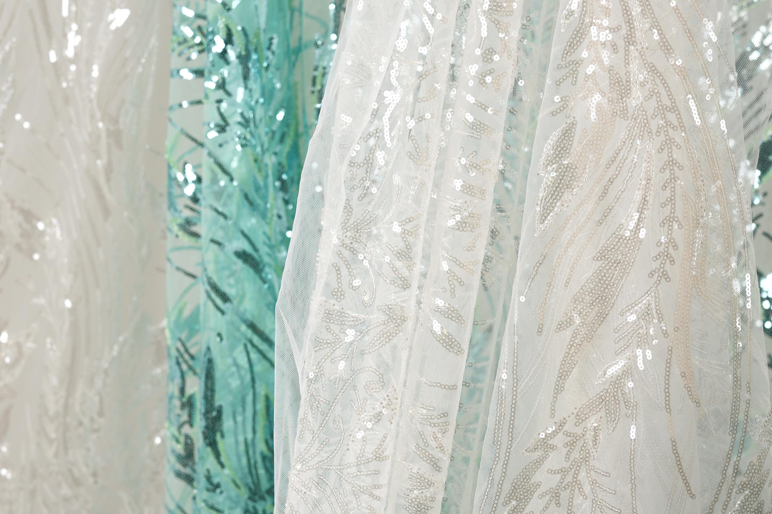 a close-up view of a white fabric with a floral pattern, as well as a fabric with a blue and green floral pattern. The fabrics appear to be made of sequins or other embellishments, adding a touch of sparkle and color to the scene.