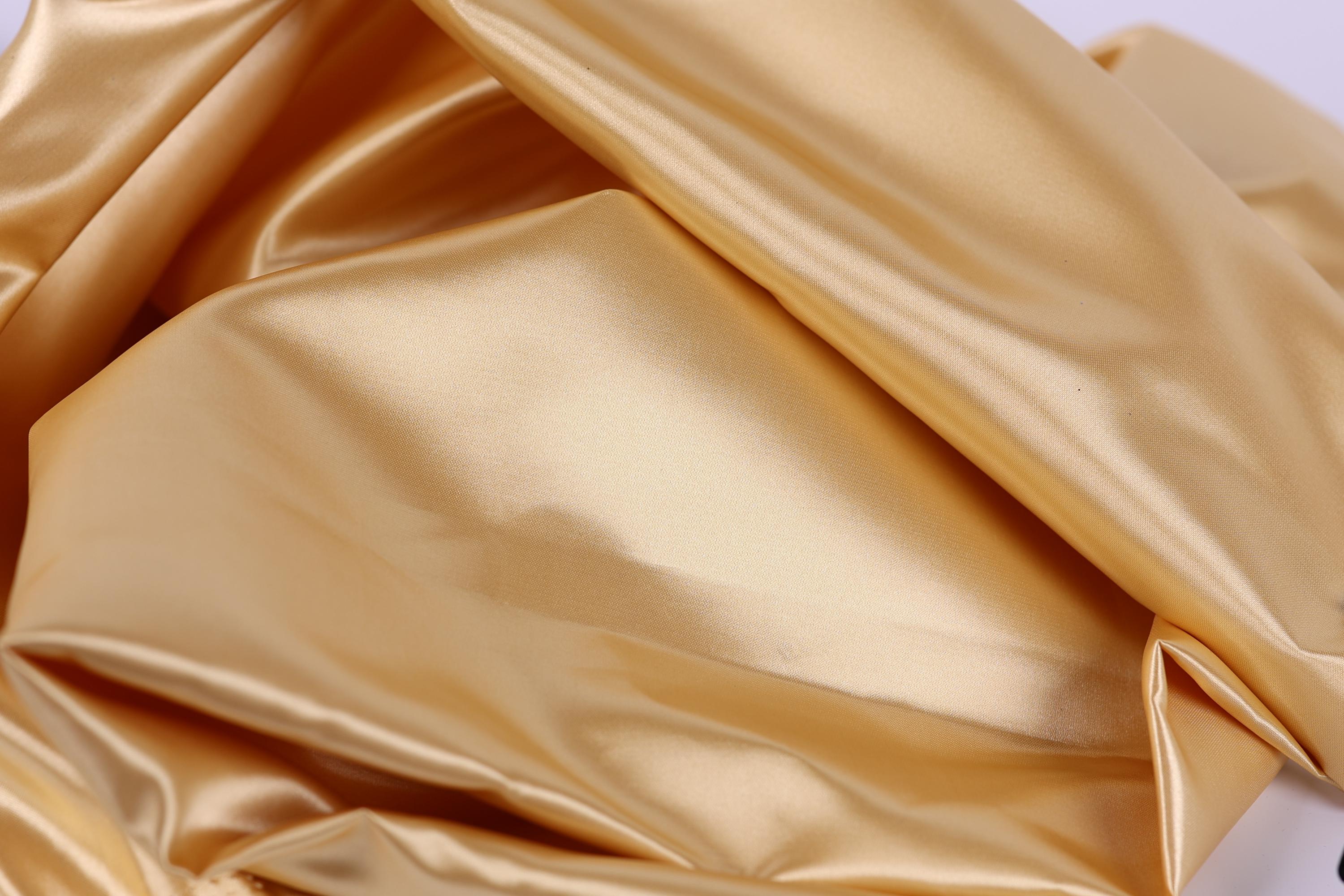 The image is a close-up of a shiny, golden-colored fabric, possibly silk or satin, with a smooth and shiny texture.