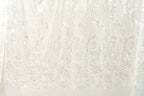 The image is a close-up of a white fabric with a repeating pattern of white leaves or flowers. The fabric appears to be made of a sheer or sheer-like material, which gives it a delicate and airy appearance. The pattern is intricate and detailed, with the leaves or flowers appearing to be made of sequins or glitter, adding a touch of sparkle to the fabric. The image does not contain any text or other discernible objects, and the focus is solely on the fabric and its pattern.